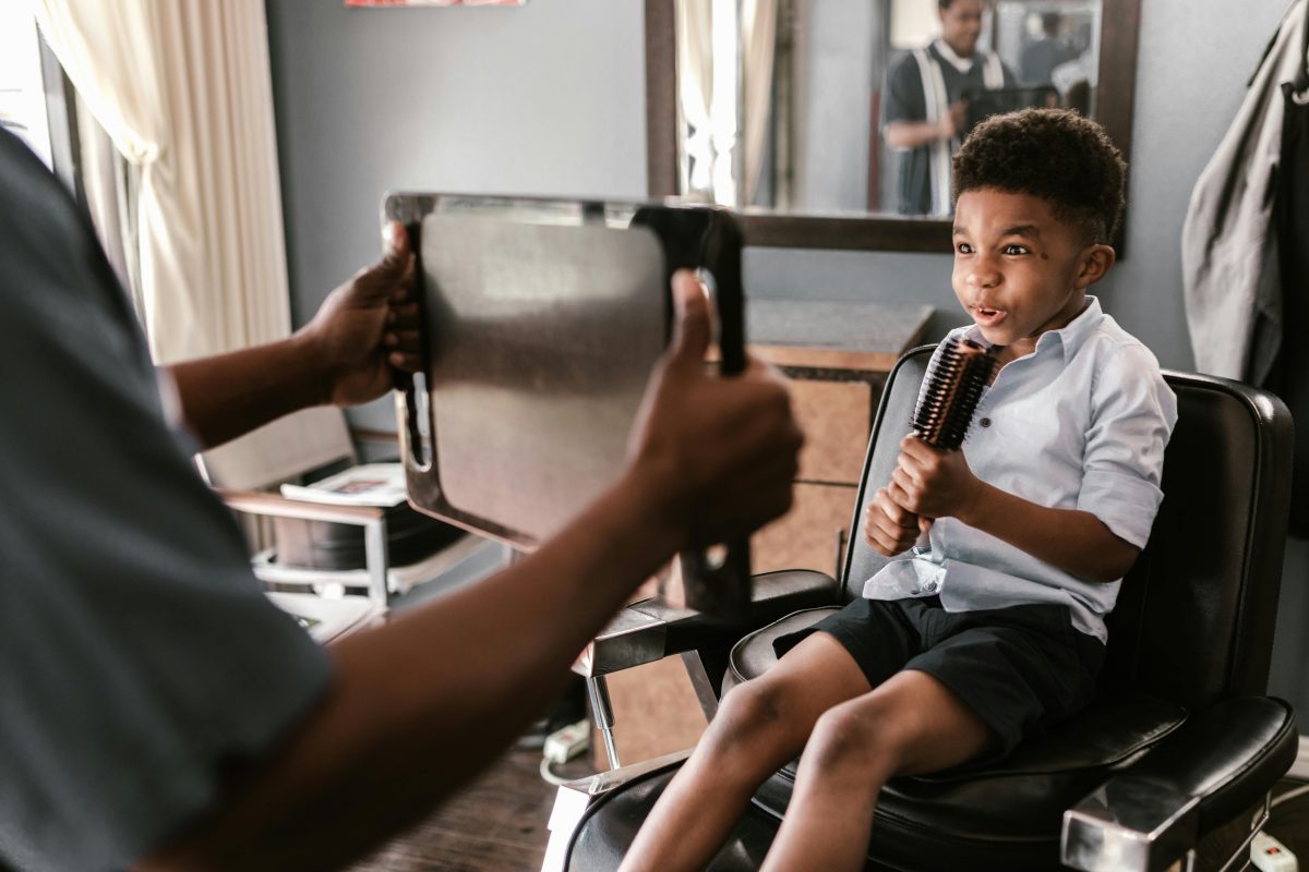 A joyful young boy sits in a barber chair holding a brush during a haircut indoors.